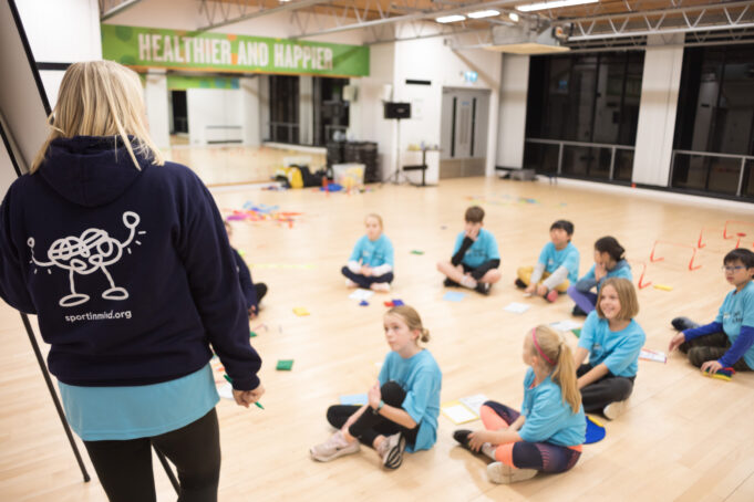 Sport in Mind A group of primary age children wearing blue tshirts sit in a circle on a wooden floor while a lady standing next to a flipchart explains something to them