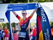 Brunnée makes history with 5th World Title A female triathlete in German kit smiles while holding a blue finish tape above her head