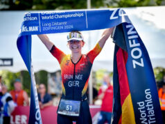 Brunnée makes history with 5th World Title A female triathlete in German kit smiles while holding a blue finish tape above her head