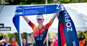 A female triathlete in German kit smiles while holding a blue finish tape above her head