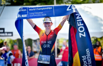 Brunnée makes history with 5th World Title A female triathlete in German kit smiles while holding a blue finish tape above her head
