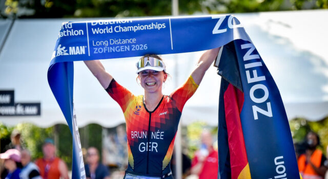Brunnée makes history with 5th World Title A female triathlete in German kit smiles while holding a blue finish tape above her head