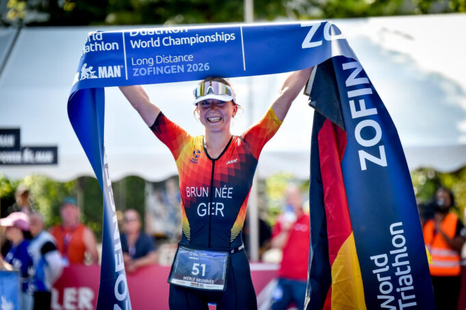 Brunnee A female triathlete in German kit smiles while holding a blue finish tape above her head