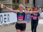City spirit shines at Run Norwich Lady in black vest and shorts makes aeroplane shape as she runs towards camera. She is smiling with her eyes closed.