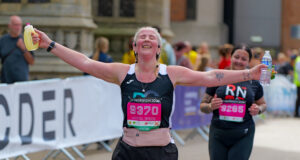 Lady in black vest and shorts makes aeroplane shape as she runs towards camera. She is smiling with her eyes closed.