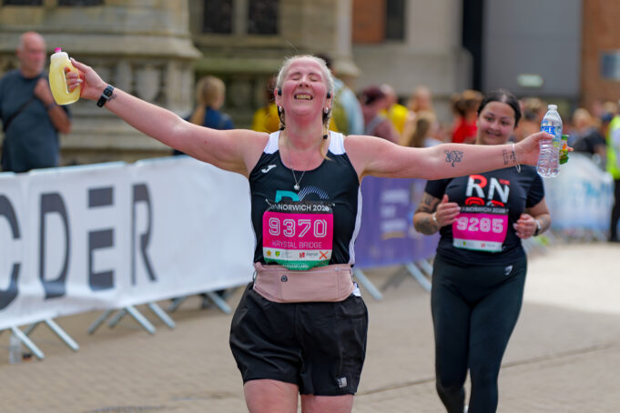 Lady in black vest and shorts makes aeroplane shape as she runs towards camera. She is smiling with her eyes closed.