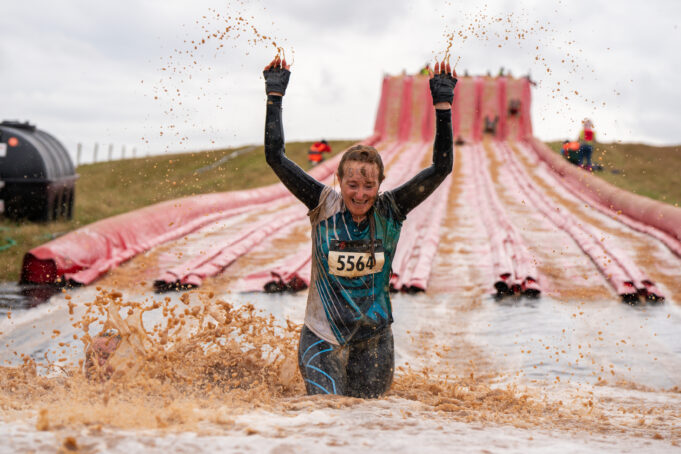 A very muddy runner splashes through a muddy thigh deep puddle at the bottom of an inflatable slide