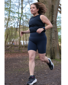 A woman runs through woods wearing navy shorts and a black vest and running shoes. Her dark crown hair is loose.