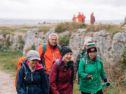OutdoorXpo welcomes The Ramblers as hiking partner 4 people of the older variety in hiking gear walk past a stone outcrop.