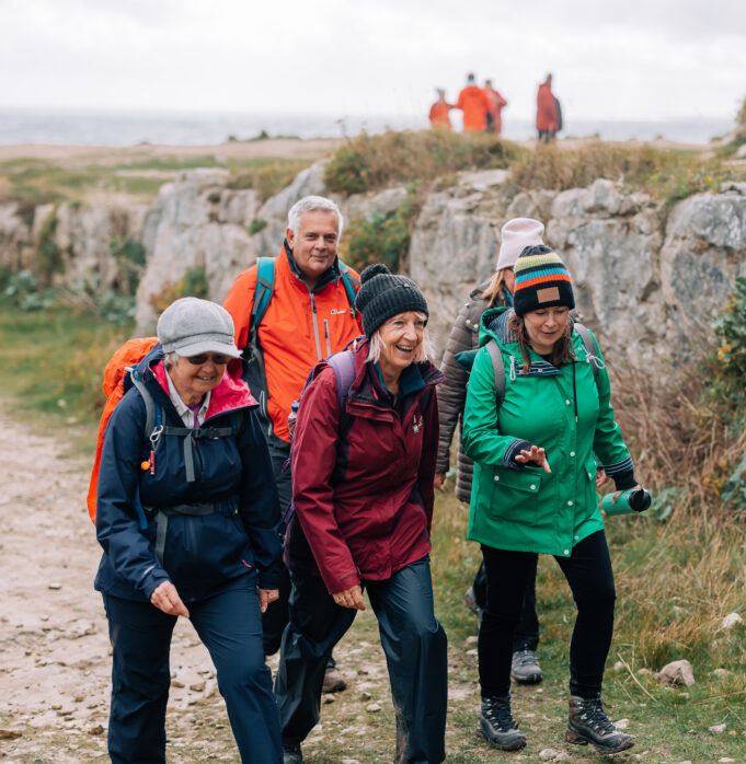 4 people of the older variety in hiking gear walk past a stone outcrop.
