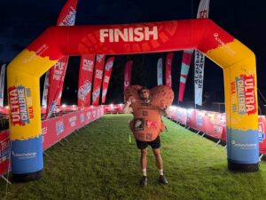 A man wearing an inflatable penis and testicles stands under a finish gantry in the dark.