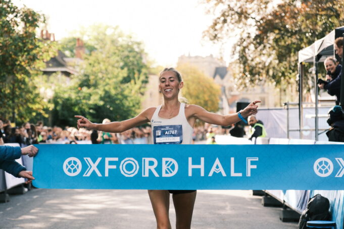 Oxford Half First female finisher Woman runs through light blue finish tape saying Oxford Half