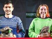 A man and woman both wearing warm jumpers and holding a dragon trophy smile for the camera