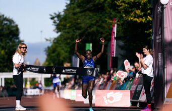 New women’s course record at Cardiff Half Woman runs through finish tape, her hands above her head, smiling.