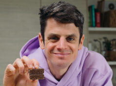 Jonny Brownlee joins One Good Thing to eliminate plastic waste Jonny Brownlee sits in a kitchen wearing a lilac hoody. He's holding a small square brown snack in his hand.
