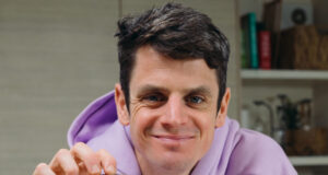 Jonny Brownlee sits in a kitchen wearing a lilac hoody. He's holding a small square brown snack in his hand.