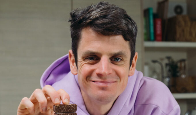 Jonny Brownlee joins One Good Thing to eliminate plastic waste Jonny Brownlee sits in a kitchen wearing a lilac hoody. He's holding a small square brown snack in his hand.