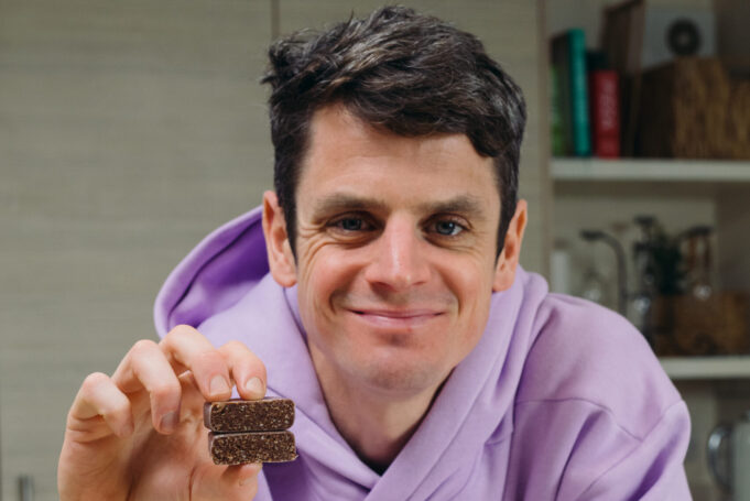jonny-brownlee-ogt-3 Jonny Brownlee sits in a kitchen wearing a lilac hoody. He's holding a small square brown snack in his hand.